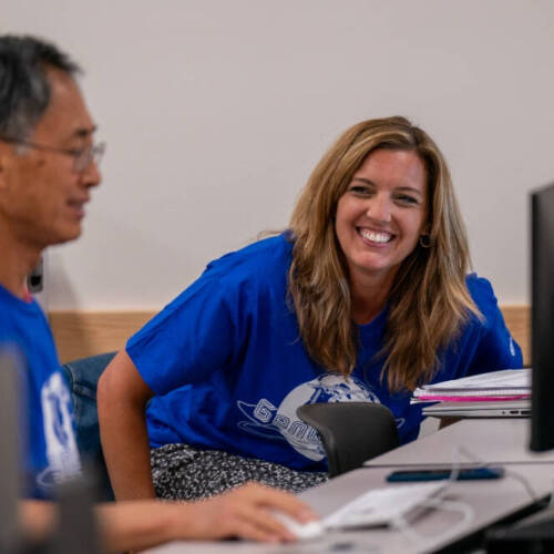Two adults in blue T-shirts smile and interact in a computer lab, with the woman in focus laughing while seated next to computer monitors.